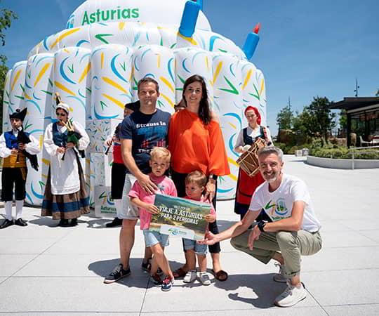 Una familia con dos niños en primer plano con la tarjeta ganadora de un premio delante de la tarta inchable conmemorativa de los 40 años de la marca Asturias paraíso natural.