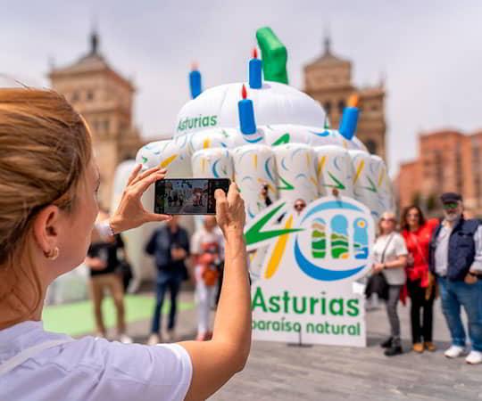 Una mujer en primer plano haciendo una fotografía con un movil a un grupo de personas delante de la tarta inchable conmemorativa de los 40 años de la marca Asturias paraíso natural.