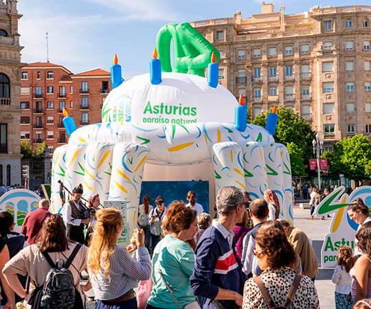 Personas en primer plano pasando por delante de la tarta inchable conmemorativa de los 40 años de la marca Asturias paraíso natural.